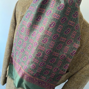 May include: A green and purple paisley patterned scarf draped over a brown tweed jacket. The scarf features a repeating design of paisley shapes and rectangular blocks in shades of green, purple, and red. The scarf's border is a solid green color.