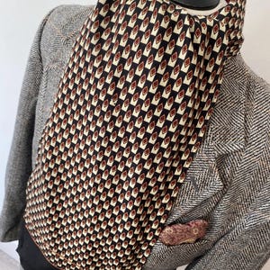 Puede incluir: Una bufanda estampada con un diseño geométrico en negro, crema y burdeos está drapeada sobre un maniquí. La bufanda se combina con una blazer de tweed gris y un pañuelo de bolsillo estampado es visible en el bolsillo de la blazer.