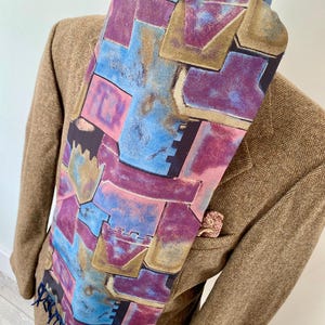 Puede incluir: Bufanda de seda colorida con un patrón geométrico en tonos azules, morados, rosas y dorados. La bufanda está drapeada sobre una blazer de tweed marrón. La bufanda tiene flecos en la parte inferior.