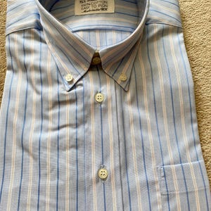 Puede incluir: Camisa azul claro con rayas verticales blancas y azules. La camisa tiene cuello abotonado, bolsillo en el pecho y una etiqueta que dice "15% St Michael 39-40 100% Cotton Easy to Iron Made in the UK."