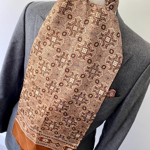 May include: A patterned silk scarf in shades of brown, tan, and cream, draped over a grey blazer. The scarf features a repeating geometric design and a solid brown border. A matching pocket square is visible in the blazer's pocket.