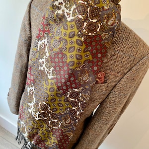 Może przedstawiać: Brązowo-zielony jedwabny szal w wzór paisley z frędzlami. Szal jest złożony i drapowany na brązowej tweedowej kurtce.