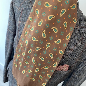 May include: A brown paisley patterned scarf with green and orange accents. The scarf is folded and displayed on a tweed jacket.