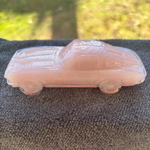 May include: A pink glass car ornament. The car is a coupe shape with visible wheels and a curved roof. The glass has an opalescent quality, giving it a soft, glowing appearance. The car is a decorative item, likely for display. The car is made of glass.