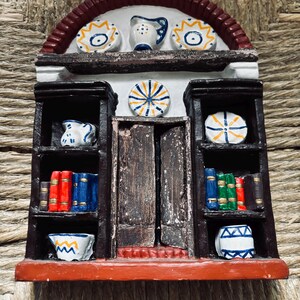 May include: A miniature ceramic bookshelf with a red arched roof, white walls, and brown shelves. The shelves are decorated with blue and white ceramic plates, pitchers, and books.