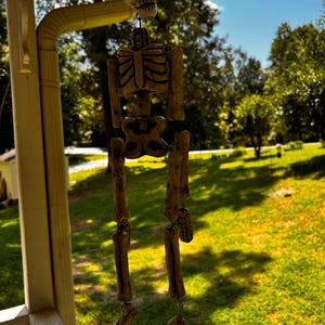 May include: A hanging skeleton decoration featuring a beige, textured skull, rib cage, pelvis, and limb bones connected by dark wire links. The figure is suspended from a structure, displaying a weathered, bone-like appearance with dark detailing.