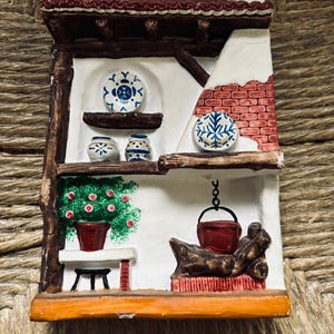 May include: A white ceramic figurine of a small house with a red brick chimney and a brown roof. The house has a white interior with blue and white painted dishes, a red pot with green plants, and a red pot hanging from a chain.