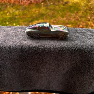 May include: A small, iridescent toy car with a sleek design. The car has a dark body with colorful highlights and sits on a gray surface. The car's wheels are visible, and the overall aesthetic suggests a vintage or collectible item.