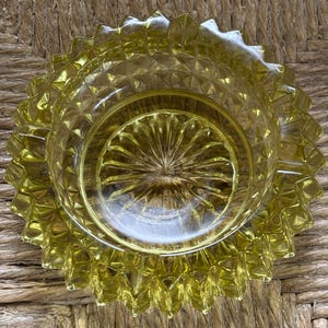 以下が含まれることがあります： ヴィンテージの黄色がかったガラス製灰皿。 灰皿は円形で、ダイヤモンド模様のテクスチャード加工された内部と中央にスターバーストのデザインがあります。 リムには、一連の尖った三角形のエッジがあります。 灰皿は織り込まれた表面にあります。