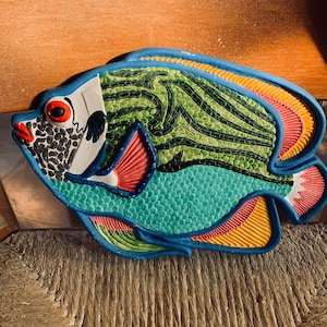 May include: A colorful ceramic fish wall decor with blue, green, yellow, red, and white paint. The fish has a stylized design with scales and a large eye.