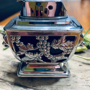May include: A silver table lighter with a grapevine design. The lighter has a square base and a rectangular top with a round lid.