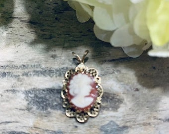 May include: A gold cameo pendant with a white cameo of a woman's profile. The cameo is set in a delicate filigree frame.