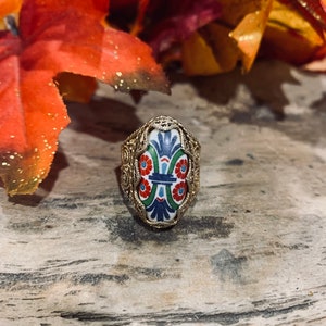 May include: A silver ring with a colorful, oval-shaped ceramic inlay featuring a floral design. The inlay has blue, green, red, and white accents.