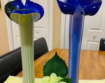 May include: Two glass vases, one with a green and blue design and the other with a blue design, are arranged with a bouquet of white flowers and green leaves.
