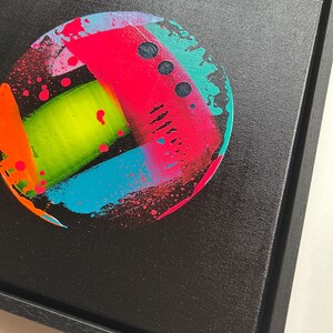 Pode incluir: Uma moldura preta quadrada contendo uma pintura circular em tela. A obra apresenta um fundo preto com uma composi&ccedil;&atilde;o abstrata vibrante de salpicos, tra&ccedil;os e pontos de tinta rosa n&eacute;on, verde-lima brilhante, turquesa e laranja.