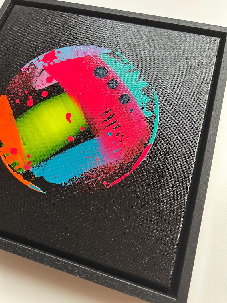 Pode incluir: Uma moldura preta quadrada contendo uma pintura circular em tela. A obra apresenta um fundo preto com uma composi&ccedil;&atilde;o abstrata vibrante de salpicos, tra&ccedil;os e pontos de tinta rosa n&eacute;on, verde-lima brilhante, turquesa e laranja.