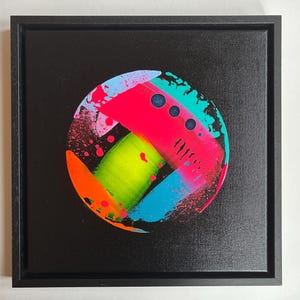 Pode incluir: Uma moldura quadrada preta contendo uma pintura abstrata circular sobre um fundo preto. O design circular apresenta pinceladas neon rosa, verde-lim&atilde;o, azul brilhante e laranja com salpicos de tinta e tr&ecirc;s pontos circulares escuros.