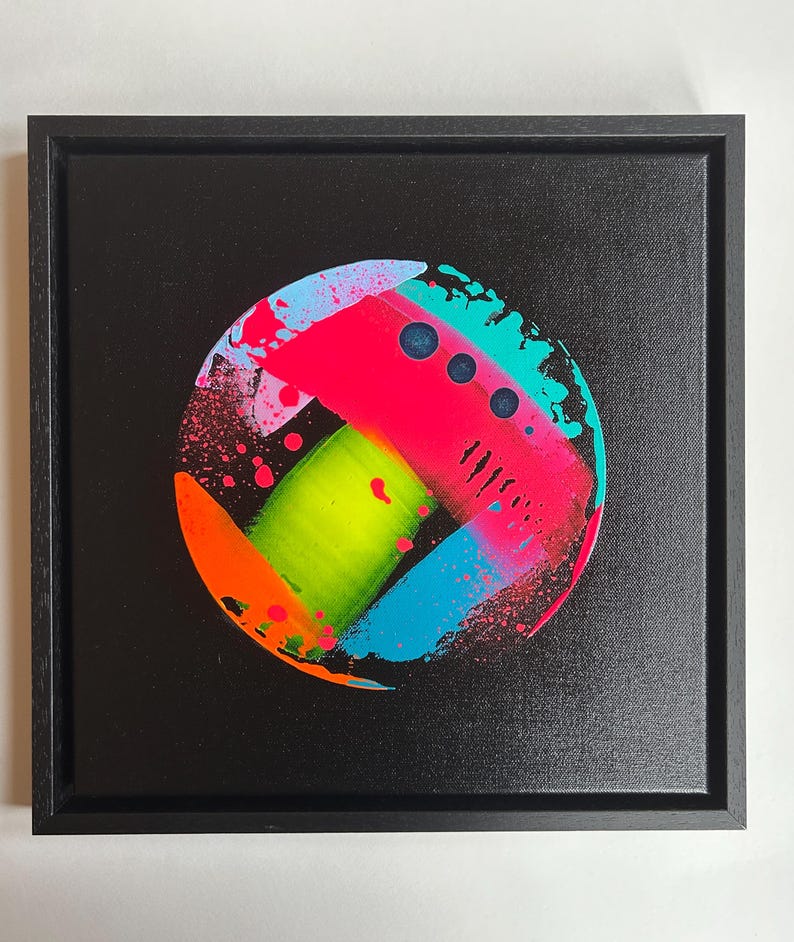Pode incluir: Uma moldura quadrada preta contendo uma pintura abstrata circular sobre um fundo preto. O design circular apresenta pinceladas neon rosa, verde-lim&atilde;o, azul brilhante e laranja com salpicos de tinta e tr&ecirc;s pontos circulares escuros.