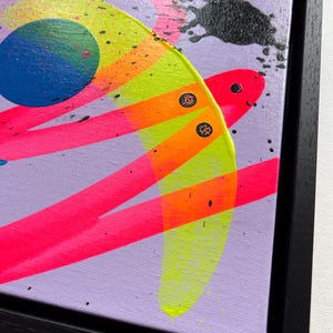 Puede incluir: Una pintura abstracta sobre lienzo en un marco negro que presenta un fondo morado claro con pinceladas de color rosa, naranja y amarillo ne&oacute;n, un c&iacute;rculo azul s&oacute;lido y salpicaduras de pintura negra con dos peque&ntilde;os detalles circulares.