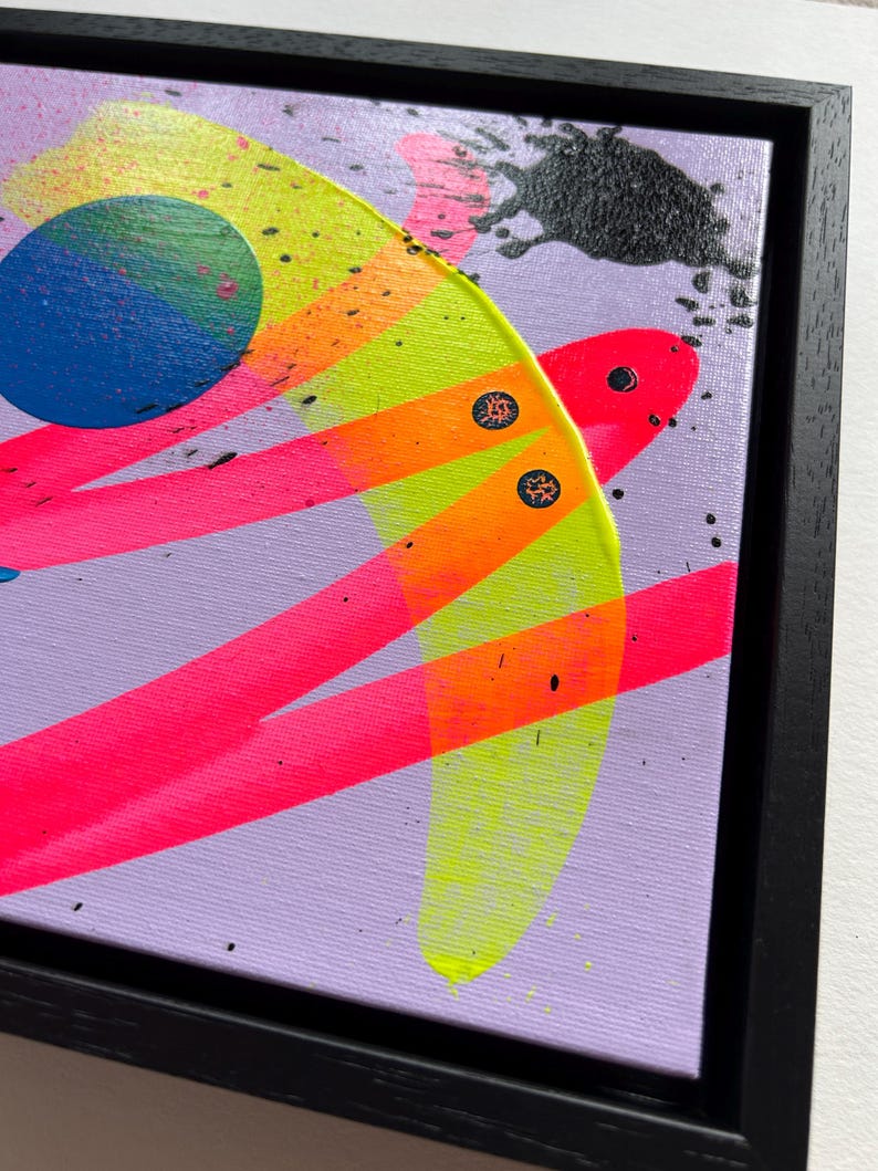 Puede incluir: Una pintura abstracta sobre lienzo en un marco negro que presenta un fondo morado claro con pinceladas de color rosa, naranja y amarillo ne&oacute;n, un c&iacute;rculo azul s&oacute;lido y salpicaduras de pintura negra con dos peque&ntilde;os detalles circulares.