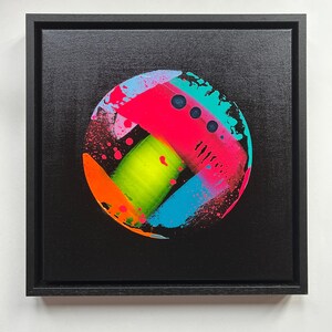 Pode incluir: Uma pintura em tela quadrada numa moldura preta apresentando uma composi&ccedil;&atilde;o circular sobre um fundo preto. O c&iacute;rculo cont&eacute;m salpicos e pinceladas vibrantes de rosa n&eacute;on, verde lima, laranja brilhante e azul ciano com pequenos pontos escuros.