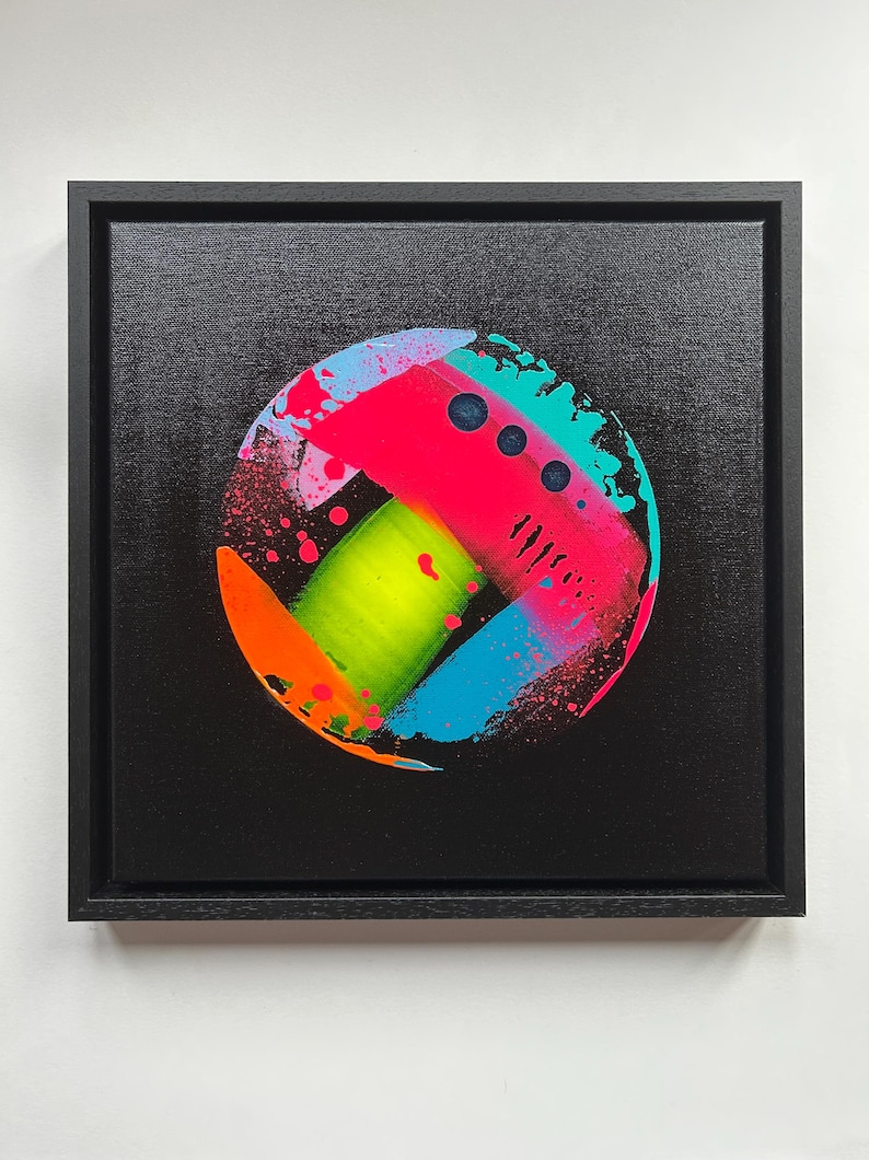 Pode incluir: Uma pintura em tela quadrada numa moldura preta apresentando uma composi&ccedil;&atilde;o circular sobre um fundo preto. O c&iacute;rculo cont&eacute;m salpicos e pinceladas vibrantes de rosa n&eacute;on, verde lima, laranja brilhante e azul ciano com pequenos pontos escuros.
