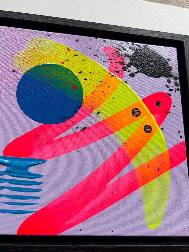 Puede incluir: Una pintura abstracta sobre lienzo en un marco negro con fondo lavanda, pinceladas en rosa y amarillo ne&oacute;n, un c&iacute;rculo azul s&oacute;lido, una forma horizontal azul texturizada y salpicaduras de pintura negra sobre la superficie.