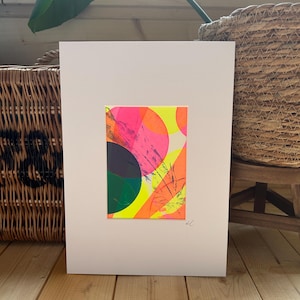 May include: Abstract art print with a colourful design of overlapping circles and brushstrokes in neon pink, orange, yellow, green, and black. The artwork is mounted on a white mount, creating a striking contrast. The piece is signed in the lower right corner.