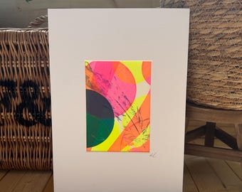 Puede incluir: Impresión artística abstracta con una vibrante composición de círculos superpuestos y pinceladas en rosa neón, naranja, amarillo, verde y negro. La obra está montada sobre un passe-partout blanco, creando un contraste llamativo. La pieza está firmada en la esquina inferior derecha.