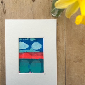 May include: Abstract art print in a white mount. The artwork features a blue top section with white cloud-like shapes, a red middle section, and a teal bottom section. The artwork is signed in the bottom right corner. The background is a wooden surface.