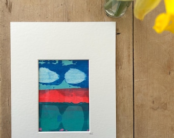 Pode incluir: Impressão de arte abstrata em um passe-partout branco. A obra de arte apresenta uma seção superior azul com formas brancas semelhantes a nuvens, uma seção média vermelha e uma seção inferior verde-azulado. A obra de arte é assinada no canto inferior direito. Fundo de madeira.