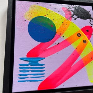 Puede incluir: Una pintura sobre lienzo cuadrada en un marco negro con un fondo violeta claro y formas abstractas. Incluye un c&iacute;rculo azul, pinceladas de color rosa ne&oacute;n y amarillo, un elemento de rayas horizontales azules y salpicaduras de pintura negra.