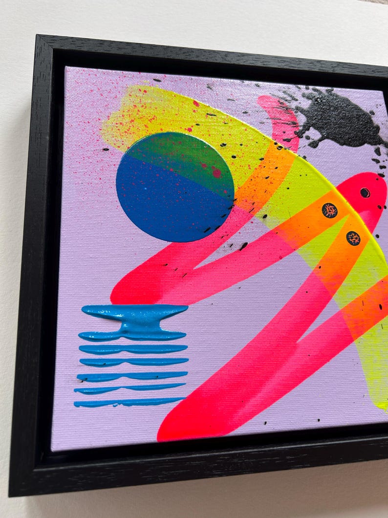 Puede incluir: Una pintura sobre lienzo cuadrada en un marco negro con un fondo violeta claro y formas abstractas. Incluye un c&iacute;rculo azul, pinceladas de color rosa ne&oacute;n y amarillo, un elemento de rayas horizontales azules y salpicaduras de pintura negra.