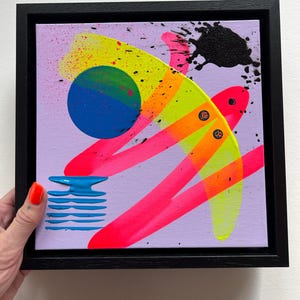 Puede incluir: Una pintura sobre lienzo cuadrada en un marco negro con fondo morado claro, un c&iacute;rculo azul grande, pinceladas en rosa ne&oacute;n y amarillo brillante, un patr&oacute;n de l&iacute;neas horizontales azules texturizadas y una salpicadura de pintura negra.