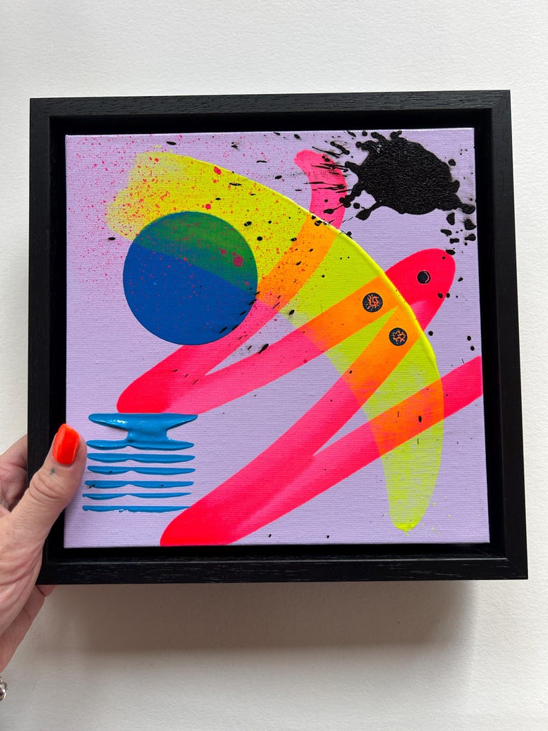Puede incluir: Una pintura sobre lienzo cuadrada en un marco negro con fondo morado claro, un c&iacute;rculo azul grande, pinceladas en rosa ne&oacute;n y amarillo brillante, un patr&oacute;n de l&iacute;neas horizontales azules texturizadas y una salpicadura de pintura negra.