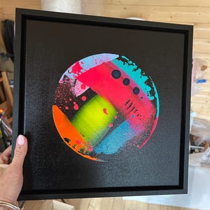 Pode incluir: Uma tela quadrada preta emoldurada em preto, apresentando uma pintura abstrata circular no centro. O design inclui salpicos de n&eacute;on vibrantes em rosa, verde-lim&atilde;o, laranja brilhante e azul ciano, com tr&ecirc;s pequenos c&iacute;rculos escuros alinhados.