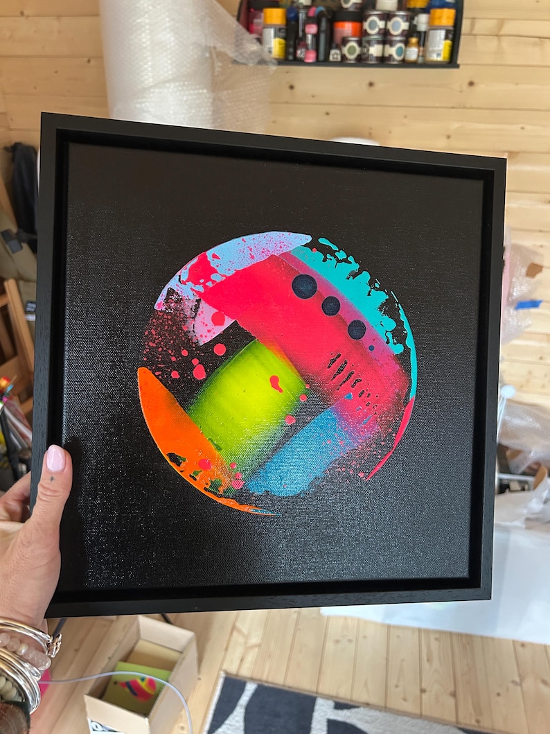 Pode incluir: Uma tela quadrada preta emoldurada em preto, apresentando uma pintura abstrata circular no centro. O design inclui salpicos de n&eacute;on vibrantes em rosa, verde-lim&atilde;o, laranja brilhante e azul ciano, com tr&ecirc;s pequenos c&iacute;rculos escuros alinhados.