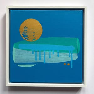 May include: A square canvas painting in a white frame featuring a solid blue background. A large gold circle sits above a horizontal block of teal and light blue paint with visible drips. Two small gold dots are positioned below the paint block.