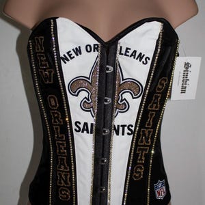 May include: Black and white corset with gold glitter and rhinestones. The corset features the New Orleans Saints logo and the words "New Orleans Saints".