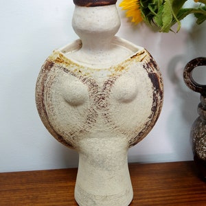May include: A cream-coloured ceramic vase in a unique, stylised human form. The vase has a rounded body with brown textured detailing and a cylindrical neck. The rim is a darker brown. A sunflower is visible in the background.