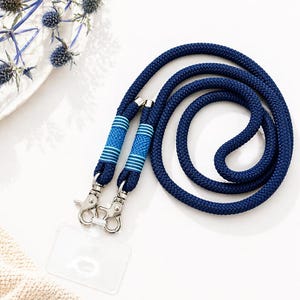 May include: A navy blue lanyard with a braided rope design. The lanyard has two blue and light blue striped accents near the metal clasps. A clear plastic ID holder is attached. The lanyard is on a white surface.