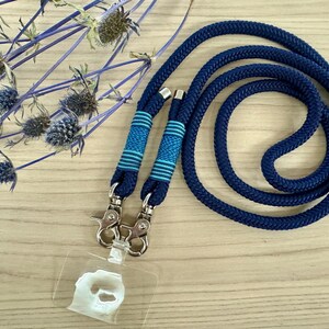 May include: A navy blue lanyard with a matching cord, featuring two sections with blue and turquoise striped accents. The lanyard has silver-colored metal clasps and a clear plastic ID holder. Decorative blue flowers are in the background.