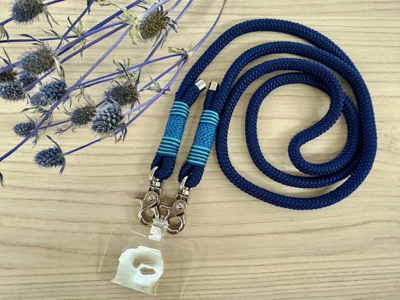 May include: A navy blue lanyard with a matching cord, featuring two sections with blue and turquoise striped accents. The lanyard has silver-colored metal clasps and a clear plastic ID holder. Decorative blue flowers are in the background.