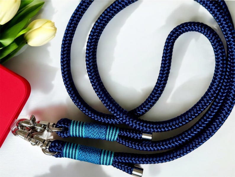 May include: A navy blue rope phone strap with silver-tone metal clasps. The strap features light blue and teal stripes near the clasps. The image also shows a red phone case and a few yellow tulips.