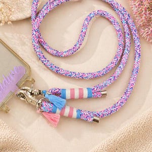 Könnte beinhalten: Ein geflochtener Handyriemen in Rosa, Blau und Lila, mit silberfarbenen Verschlüssen und Quasten. Der Riemen ist an einer transparenten Handyhülle mit dem Namen "Nadine" in Lavendelschrift befestigt. Der Riemen ist ca. 150 cm lang.