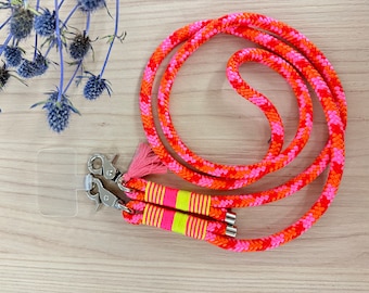 Può includere: Un vivace cordino per telefono con un design a corda intrecciata nei toni dell'arancione e del rosa. Il cordino presenta chiusure in metallo argentato, un nappa rosa e dettagli giallo neon e rosa. È visibile anche una custodia trasparente per telefono.