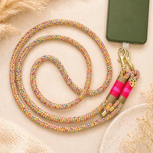 Könnte beinhalten: Ein farbenfrohes Telefon-Lanyard mit einem geflochtenen Regenbogen-Kordel. Das Lanyard hat goldfarbene Verschlüsse und rosa und braune Akzente. Eine grüne Handyhülle ist am Lanyard befestigt.