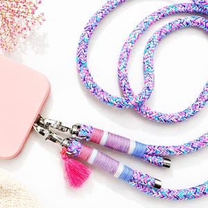 Könnte beinhalten: Ein rosa Handyhülle mit einem mehrfarbigen geflochtenen Handyband. Das Band hat rosa, blaue und violette Farbtöne, mit silbernen Metallverschlüssen und einer rosa Quaste. Das Band ist zum Tragen um den Hals oder die Schulter konzipiert.