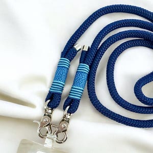 May include: A navy blue lanyard with a thick, braided rope design. The lanyard features two silver-toned metal clasps and is accented with light blue and navy blue striped details. The lanyard is designed to hold an ID badge or other small items.