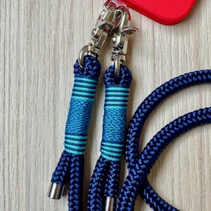 May include: Close-up of a navy blue phone lanyard with silver-tone metal clasps. The lanyard features a braided rope design with teal and blue striped accents. A red phone case is visible in the background.